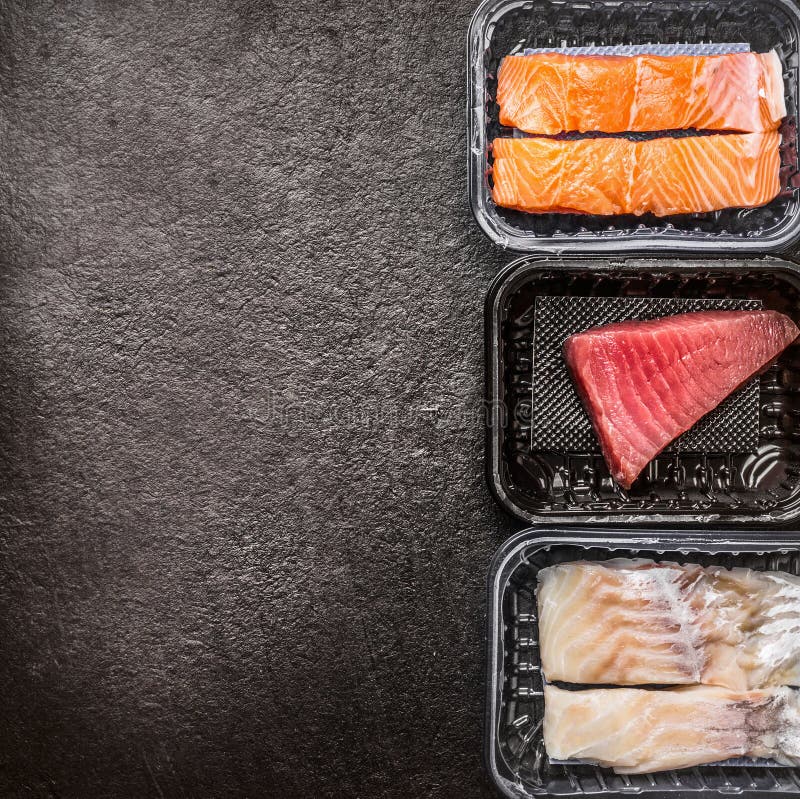 Various of raw fish fillets : salmon, tuna and codfish in plastic boxes on dark rustic background, top view, border, place for tex