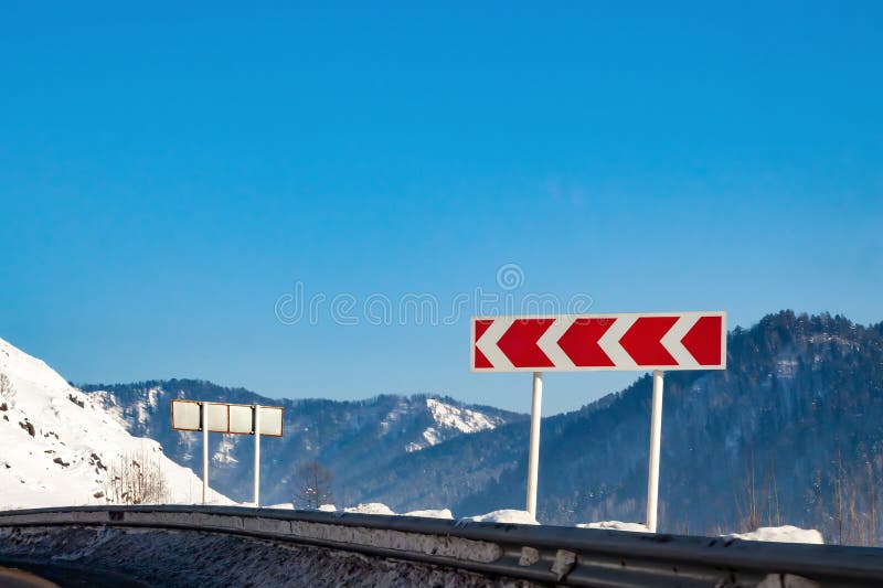 Varning för vägskyltning för en skarp och farlig vändning till vänster med röda pilar mot en vit bakgrund med berg, snö på vinter fotografering för bildbyråer Varning för vägskyltning för en skarp och farlig vändning till vänster med röda pilar mot en vit bakgrund med berg, snö på vinter fotografering för bildbyråer