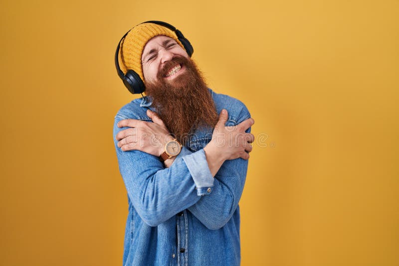 Varsam man med långt skägg som lyssnar på musik med hörlurar som kramar sig själv, glad och positiv leende tillitsfull royaltyfri fotografi