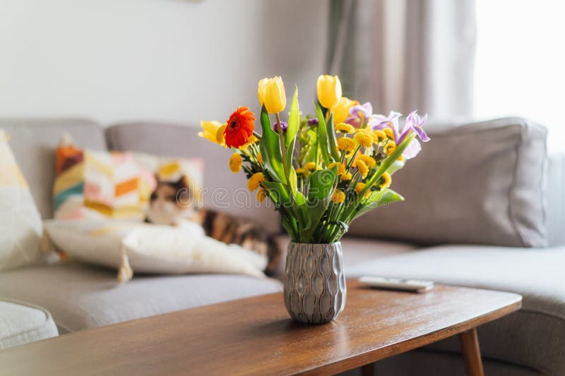 Vase of multicolor flowers on coffee table with blurred background of modern cozy living room with gray couch sofa and relaxing cat pet. Open space home interior design. Cozy home mood. Copy space.