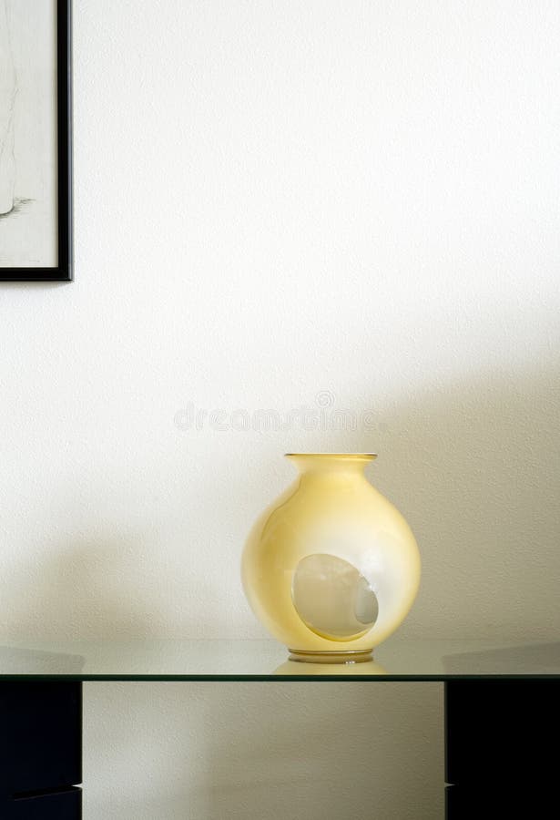 Stunning hand blown yellow and white art deco glass vase on a glass shelf in front of a rooms wall under a partial black frame drawing. Stunning hand blown yellow and white art deco glass vase on a glass shelf in front of a rooms wall under a partial black frame drawing