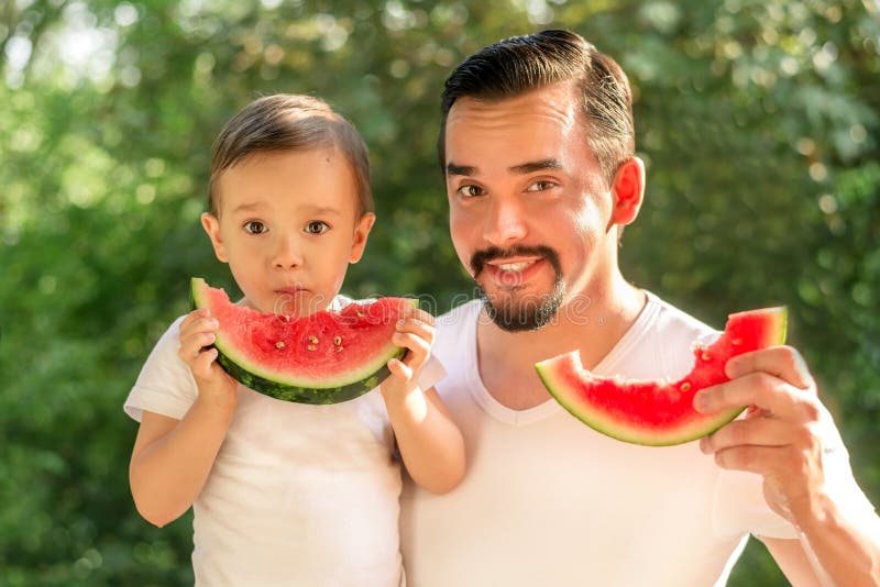 Vater und Sohn essen zusammen Wassermelonen, Mann und Kind halten Scheiben von saftigen Wassermelonen, grüne Blätter im Hintergru