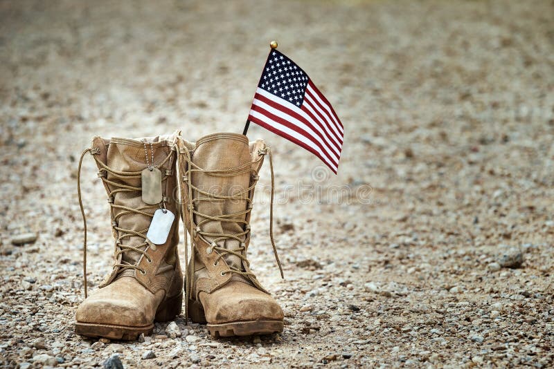 Old military combat boots with dog tags and a small American flag. Rocky gravel background with copy space. Memorial Day, Veterans day, sacrifice concept. Old military combat boots with dog tags and a small American flag. Rocky gravel background with copy space. Memorial Day, Veterans day, sacrifice concept.