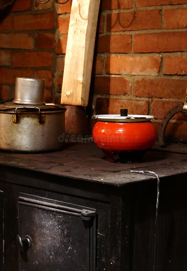On a black cast-iron stove against a brick wall stands a saucepan with a ladle and red cast iron; in the kitchen of a country house firewood is drying against the wall. On a black cast-iron stove against a brick wall stands a saucepan with a ladle and red cast iron; in the kitchen of a country house firewood is drying against the wall.