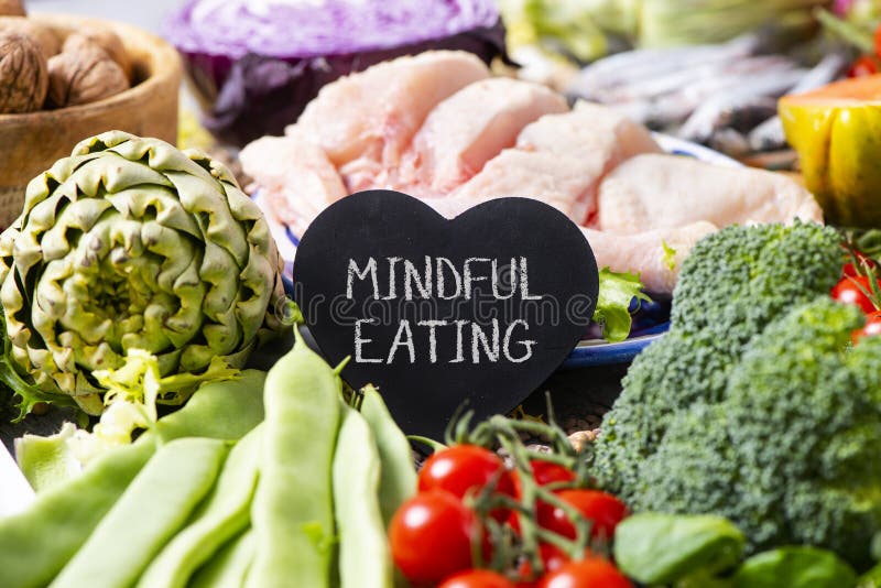 A black heart-shaped signboard with the text mindful eating, on a pile of different vegetables, such as French beans, cherry tomatoes, a head of broccoli, and some pieces of chicken in the background. A black heart-shaped signboard with the text mindful eating, on a pile of different vegetables, such as French beans, cherry tomatoes, a head of broccoli, and some pieces of chicken in the background