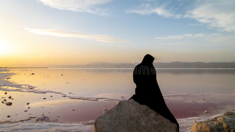 Veiled Iranian woman in hijab black dress sitting on a rock by the Maharloo pink lake