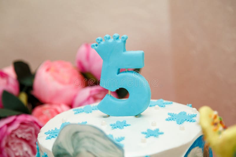 Blue candle with number 5 and crown on top of winter birthday cake with snowflakes. Frozen cartoon character thematic party. Blue candle with number 5 and crown on top of winter birthday cake with snowflakes. Frozen cartoon character thematic party