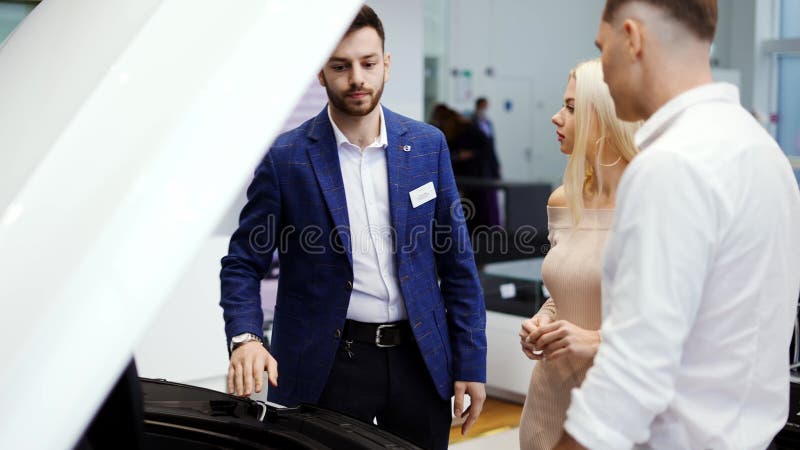 Vendedor mostrando el motor de coches en cámara abierta a pareja en cámara lenta de la sala de exposición automática almacen de metraje de vídeo