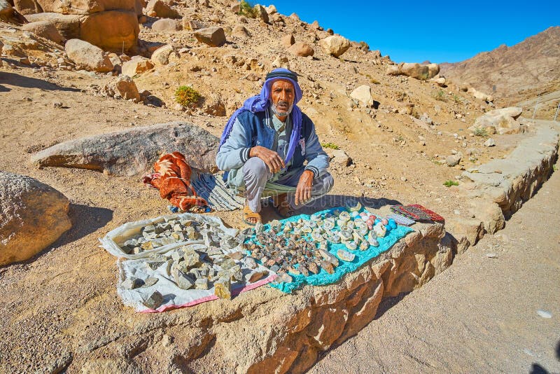 ST CATHERINE, EGYPT - DECEMBER 25, 2017: The senior bedouin vendor sits at the mountain road and sells the local souvenirs - the quartz geodes and Mount Sinai stones with plant fossils, on December 25 in St Catherine. ST CATHERINE, EGYPT - DECEMBER 25, 2017: The senior bedouin vendor sits at the mountain road and sells the local souvenirs - the quartz geodes and Mount Sinai stones with plant fossils, on December 25 in St Catherine.