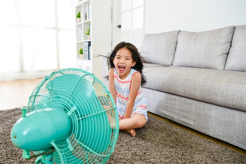 Happy attractive female kid blowing cooling wind from electric fan feeling cold looking at camera laughing in summer time. Happy attractive female kid blowing cooling wind from electric fan feeling cold looking at camera laughing in summer time.