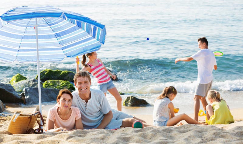 Verbinden Sie die Entspannung auf Strand während ihre Kinder, die aktive Spiele spielen stockfotos Verbinden Sie die Entspannung auf Strand während ihre Kinder, die aktive Spiele spielen stockfotos