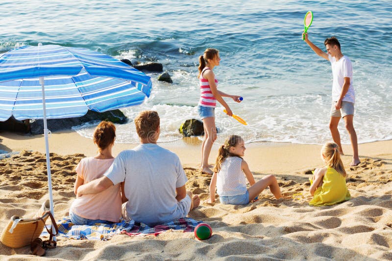 Verbinden Sie die Entspannung auf Strand während ihre Kinder, die aktive Spiele spielen stockfotos Verbinden Sie die Entspannung auf Strand während ihre Kinder, die aktive Spiele spielen stockfotos