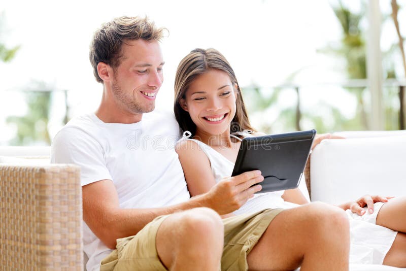 Couple relaxing together in sofa with tablet computer pc having fun. Romantic young happy multiracial couple in love sitting at home in sofa resting having fun maybe watching movie. Man and woman. Couple relaxing together in sofa with tablet computer pc having fun. Romantic young happy multiracial couple in love sitting at home in sofa resting having fun maybe watching movie. Man and woman.
