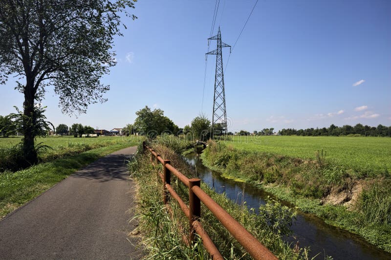 Paved path bordered by a field and a stream of water with a pylon afar on a sunny day in the italian countryside. Paved path bordered by a field and a stream of water with a pylon afar on a sunny day in the italian countryside