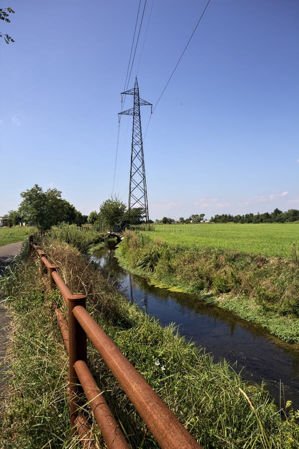 Paved path bordered by a field and a stream of water with a pylon afar on a sunny day in the italian countryside. Paved path bordered by a field and a stream of water with a pylon afar on a sunny day in the italian countryside