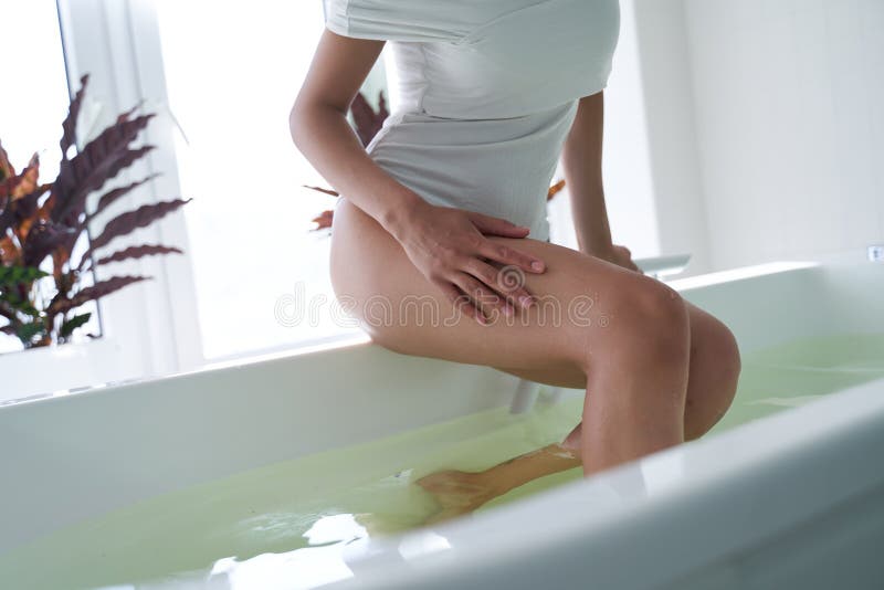 Cropped photo of unrecognized woman in a white swimsuit sitting on the edge of a bathtub with feet in the water and touching her leg. Cropped photo of unrecognized woman in a white swimsuit sitting on the edge of a bathtub with feet in the water and touching her leg