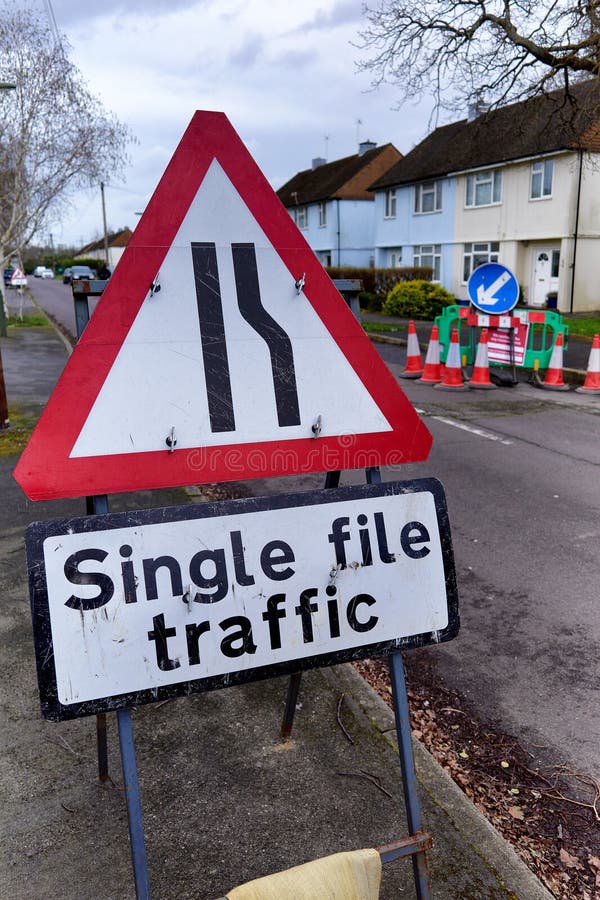 Red bordered triangular warning sign for single file traffic, at the start of roadworks on a residential road in Surrey. A rectangular sign below describes the situation and a circular blue keep left sign is to the right, in the background. Red bordered triangular warning sign for single file traffic, at the start of roadworks on a residential road in Surrey. A rectangular sign below describes the situation and a circular blue keep left sign is to the right, in the background.