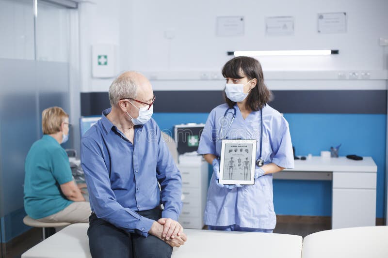Female doctor shows elderly patient wearing face mask a tablet displaying human skeleton. Digital gadget is held by nurse who is giving a medical diagnostic to senior retired man. Female doctor shows elderly patient wearing face mask a tablet displaying human skeleton. Digital gadget is held by nurse who is giving a medical diagnostic to senior retired man.