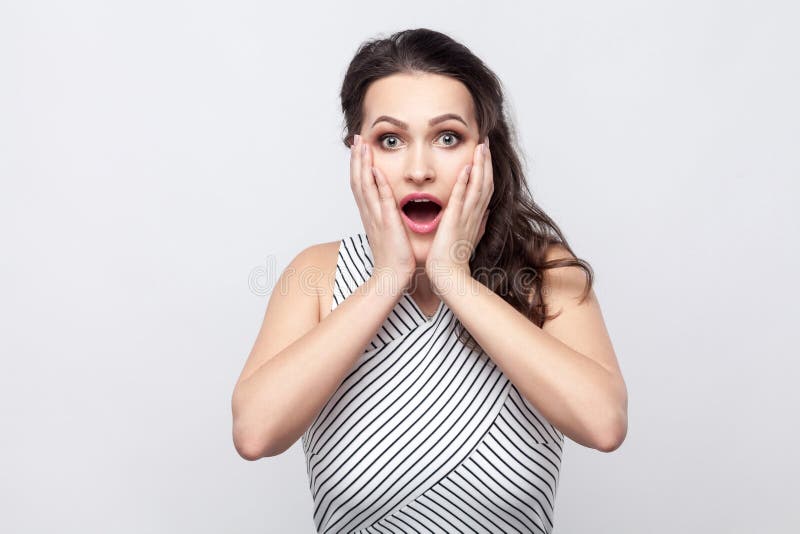 Verraste mooie jonge donkerbruine vrouw met make-up en gestreepte kleding die rakend haar gezicht en bekijkend camera met bevinde royalty-vrije stock foto's Verraste mooie jonge donkerbruine vrouw met make-up en gestreepte kleding die rakend haar gezicht en bekijkend camera met bevinde royalty-vrije stock foto's