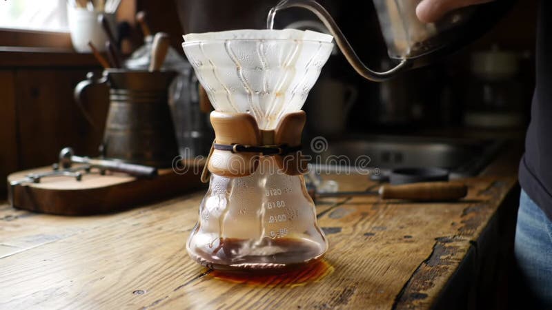 Verser de l'eau dans la machine à café Chemex sur une table en bois rustique avec des ustensiles de cuisine et un évier en arrière illustration libre de droits