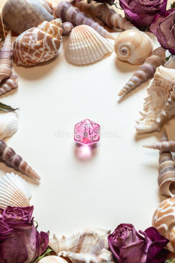 Vertical image of a transparent pink RPG gaming die on a white surface framed by seashells and dried pink roses. Vertical image of a transparent pink RPG gaming die on a white surface framed by seashells and dried pink roses