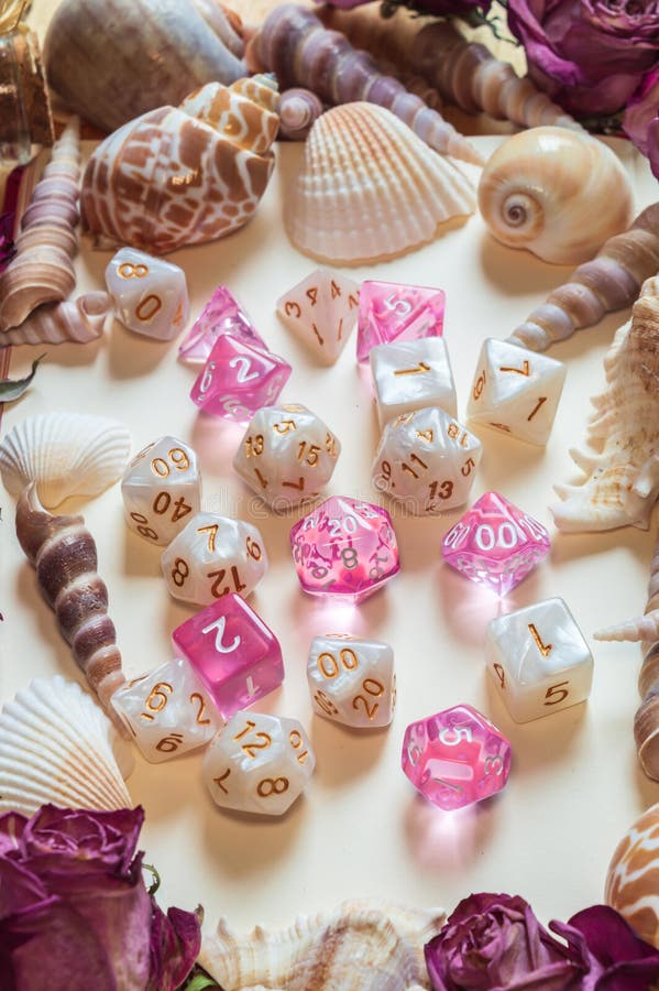 Vertical image of two sets of white and pink RPG gaming dice framed by seashells and dried pink roses. Vertical image of two sets of white and pink RPG gaming dice framed by seashells and dried pink roses