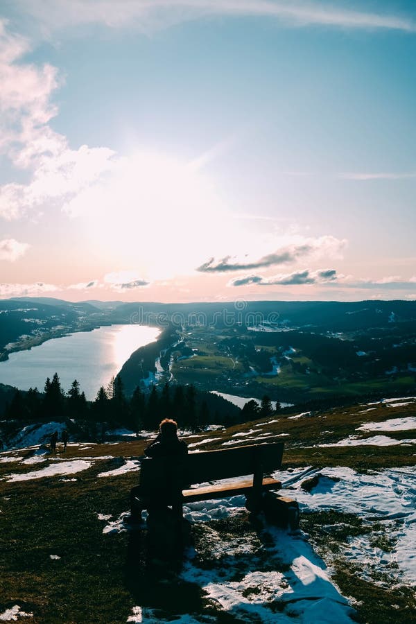 Vertical shot of a person on a bench on a snow covered hill and a beautiful lake in the background