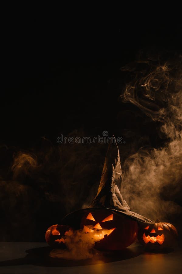 Vertikale Halloween-Karte. Hexenhut auf einem Gefäß mit geschnitzten gruseligen Grimassen auf einem schwarzen Hintergrund im Nebel stockfotografie