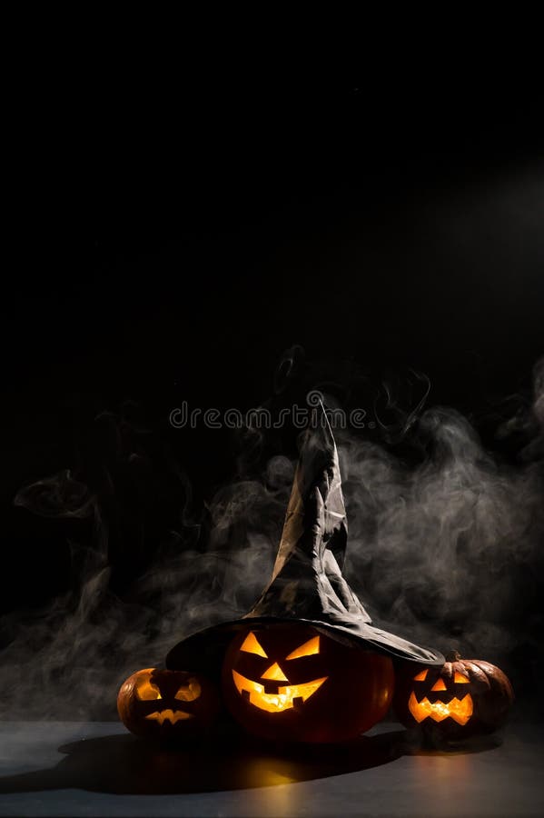 Vertikale Halloween-Karte. Hexenhut auf einem Gefäß mit geschnitzten gruseligen Grimassen auf einem schwarzen Hintergrund im Nebel lizenzfreies stockfoto