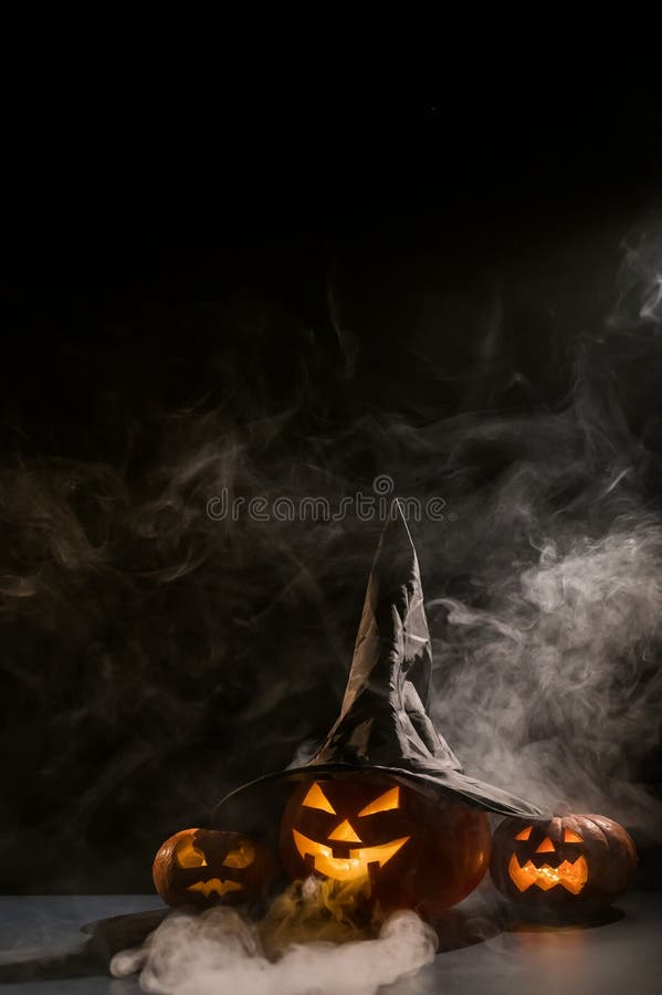 Vertikale Halloween-Karte. Hexenhut auf einem Gefäß mit geschnitzten gruseligen Grimassen auf einem schwarzen Hintergrund im Nebel stockbild