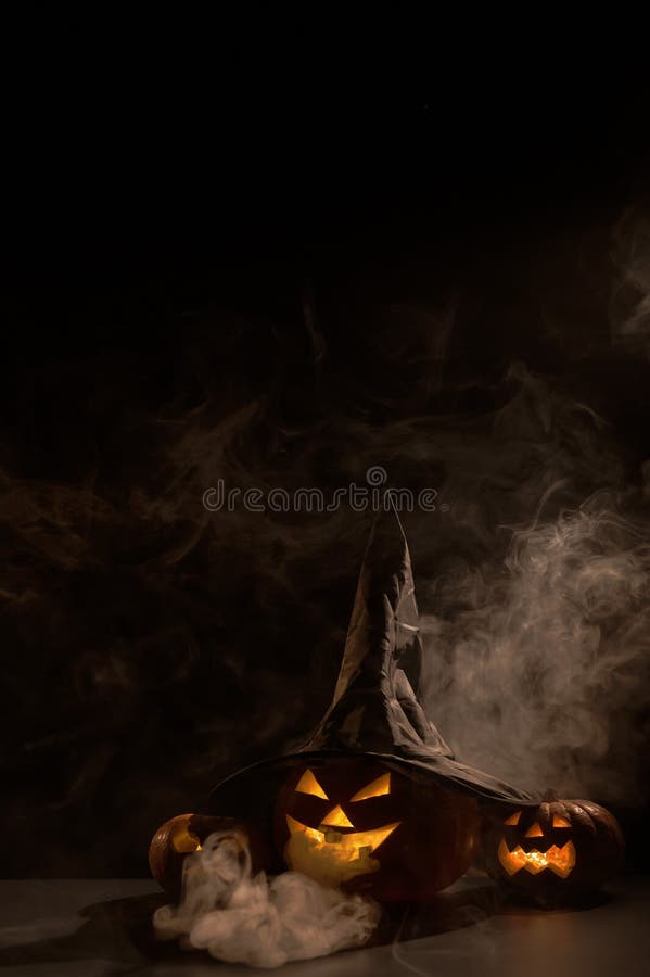 Vertikale Halloween-Karte. Hexenhut auf einem Gefäß mit geschnitzten gruseligen Grimassen auf einem schwarzen Hintergrund im Nebel stockfotografie