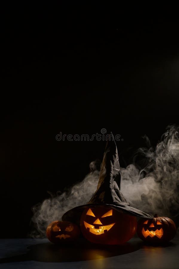 Vertikale Halloween-Karte. Hexenhut auf einem Gefäß mit geschnitzten gruseligen Grimassen auf einem schwarzen Hintergrund im Nebel stockbild