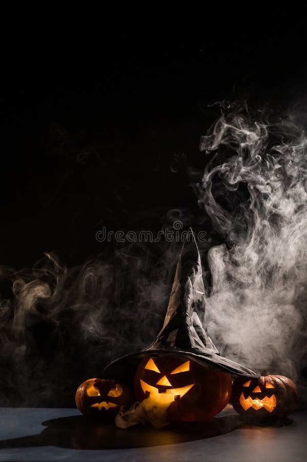 Vertikale Halloween-Karte. Hexenhut auf einem Gefäß mit geschnitzten gruseligen Grimassen auf einem schwarzen Hintergrund im Nebel stockfotografie