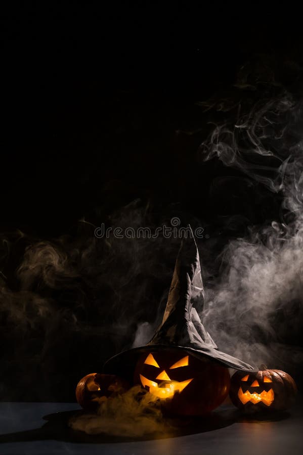 Vertikale Halloween-Karte. Hexenhut auf einem Gefäß mit geschnitzten gruseligen Grimassen auf einem schwarzen Hintergrund im Nebel stockbilder