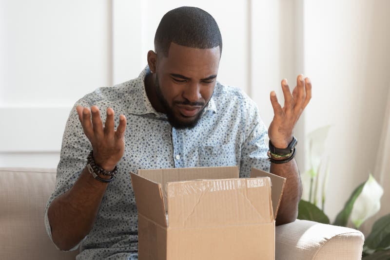 Frustrated african American millennial male buyer unpack unbox cardboard box delivery package disappointed with product quality, confused biracial man shop online open box get wrong order. Frustrated african American millennial male buyer unpack unbox cardboard box delivery package disappointed with product quality, confused biracial man shop online open box get wrong order