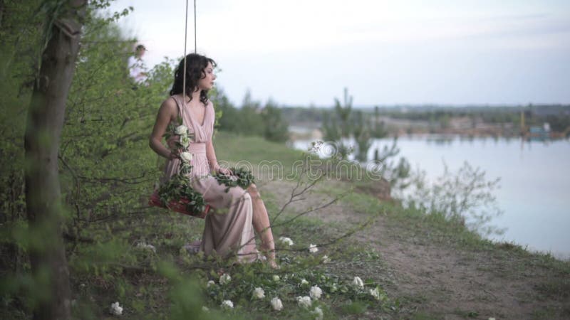 Very beautiful smiling girl in light pink dress relax on the swing decorated by flowers at the lake beach