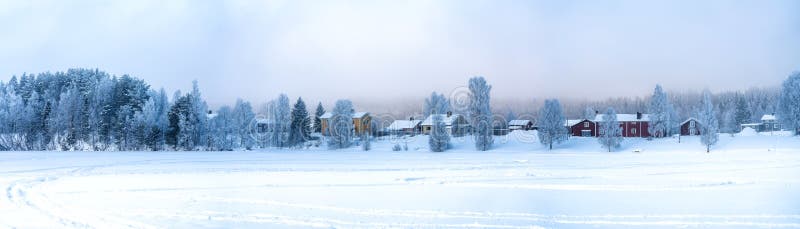 Very wide panorama view on Swedish village with red and yellow wooden houses in winter overcast on frozen river coast at spruce