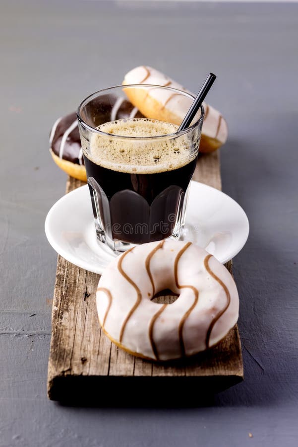 Glass of Black Americano Coffee and Tasty Chocolate and Vanilla Donuts on Wooden Tray Gray Background Morning Breakfast Lifestyle Vertical. Glass of Black Americano Coffee and Tasty Chocolate and Vanilla Donuts on Wooden Tray Gray Background Morning Breakfast Lifestyle Vertical.