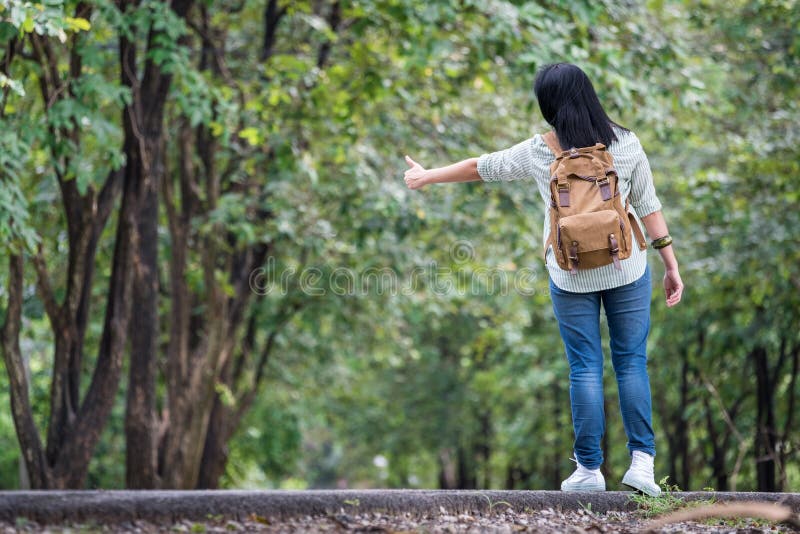 Viaggiatore con zaino e sacco a pelo asiatico della donna che sta sulla strada della campagna con l'albero dentro fotografia stock libera da diritti Viaggiatore con zaino e sacco a pelo asiatico della donna che sta sulla strada della campagna con l'albero dentro fotografia stock libera da diritti