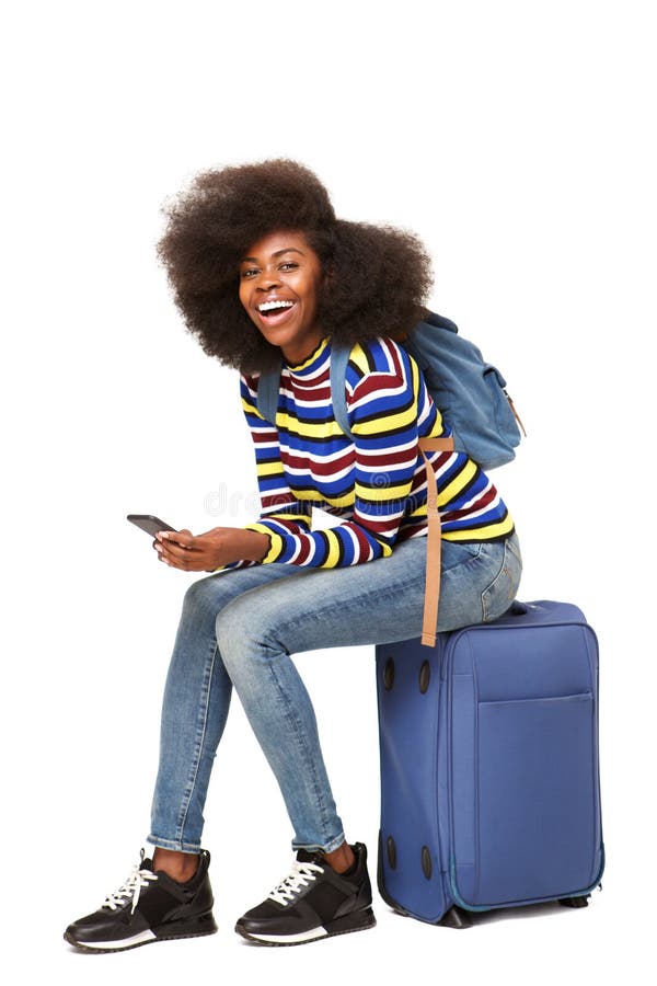 Viajero femenino afroamericano joven que se sienta en la maleta con el teléfono móvil imagenes de archivo