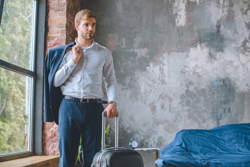Viajero masculino confiado con la chaqueta sobre la situación del hombro con la maleta en dormitorio foto de archivo libre de regalías