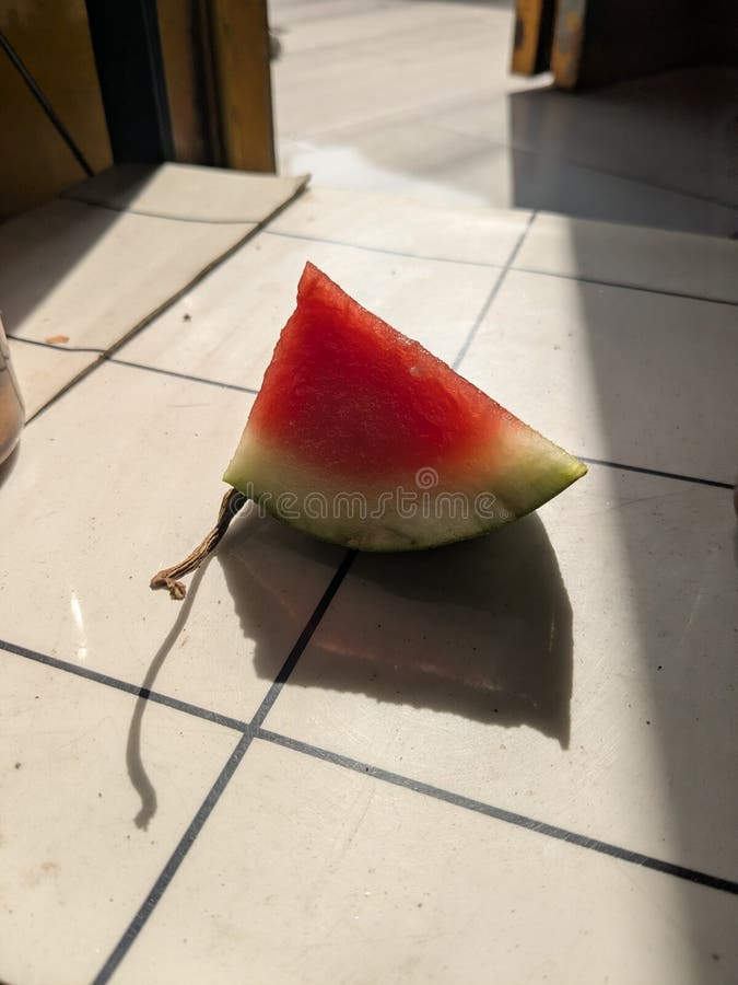 A vibrant, triangular slice of ripe red watermelon with its green rind rests on a light-colored tiled floor. Strong natural sunlight casts dramatic shadows, including a distinct shadow from the watermelon itself and its small, dried stem. The simple composition highlights the refreshing fruit, evoking feelings of summer and healthy eating. Perfect for food blogs, summer promotions, healthy lifestyle content, or any project needing a fresh, natural fruit image with interesting light. Triangular cut watermelon stock images, royalty-free photos and pictures