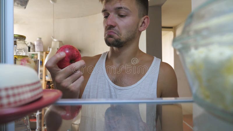 Vicino a un uomo assonnato che cerca un cibo in frigo per cucinare la colazione. giovane pigro che trova la mela sulla mensola, la