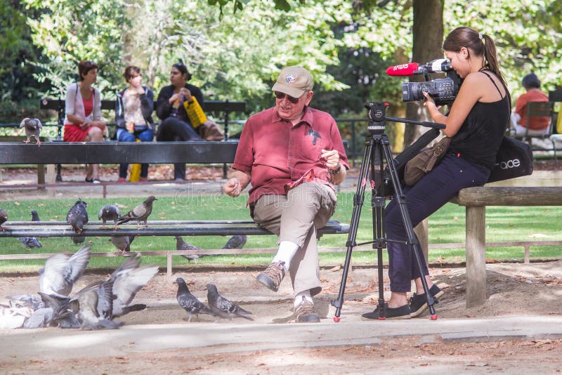 Young woman videographer films smiling older gentleman in Peace Corps hat as he feeds the pigeons in Luxembourg Garden, Paris, France. Young woman videographer films smiling older gentleman in Peace Corps hat as he feeds the pigeons in Luxembourg Garden, Paris, France