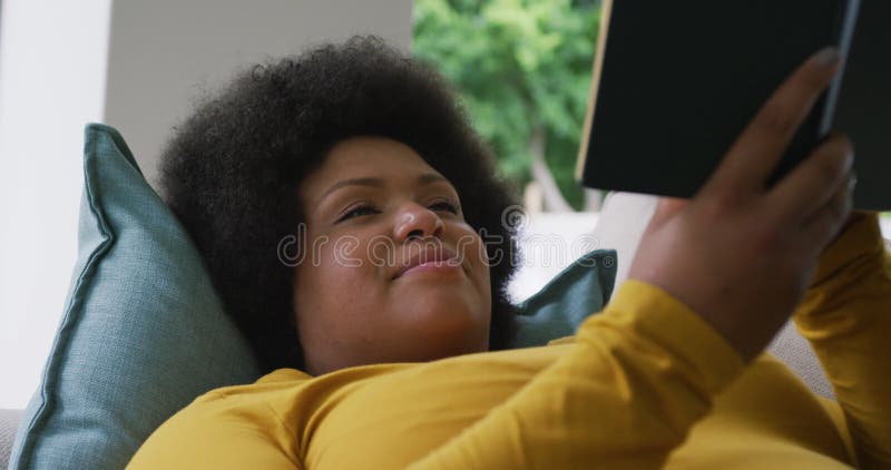 Vidéo d'une femme afro-américaine de taille plus heureuse allongée sur un canapé et le livre de lecture clips vidéos