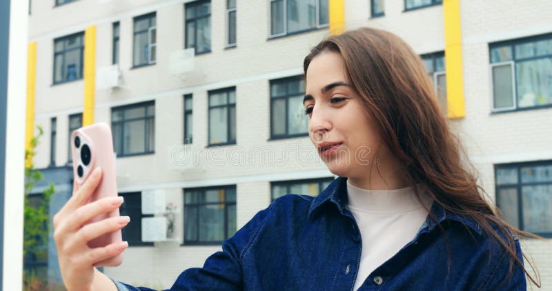Vidéo d'une jeune femme habillée en cascade tenant un smartphone près du visage debout devant le bâtiment résidentiel clips vidéos