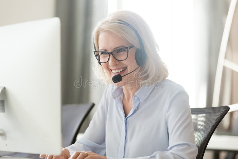 Vieja empresaria feliz en las auriculares que hacen la llamada que mira el ordenador