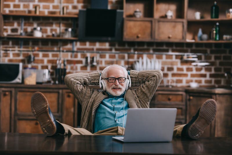 Viejo hombre relajado en auriculares usando el ordenador portátil con los pies