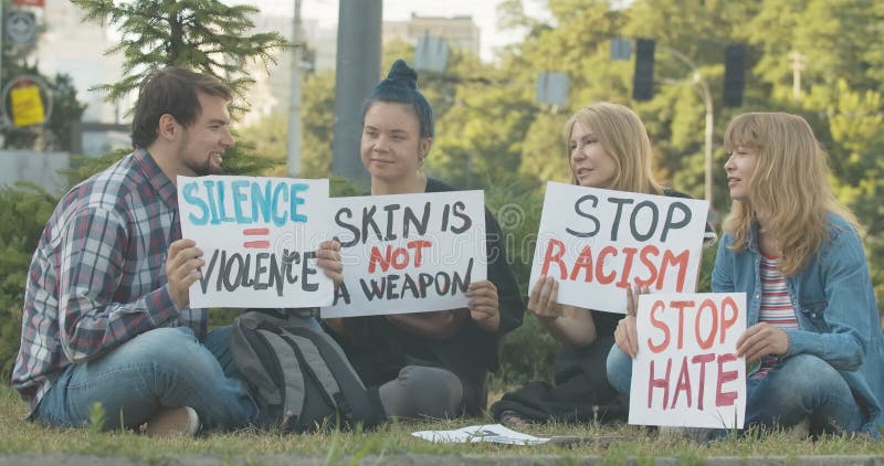 Vier kaukasische demonstranten zitten op gras met een spandoek tegen racisme en spreken. groep van caucasiaanse mannen en vrouwen