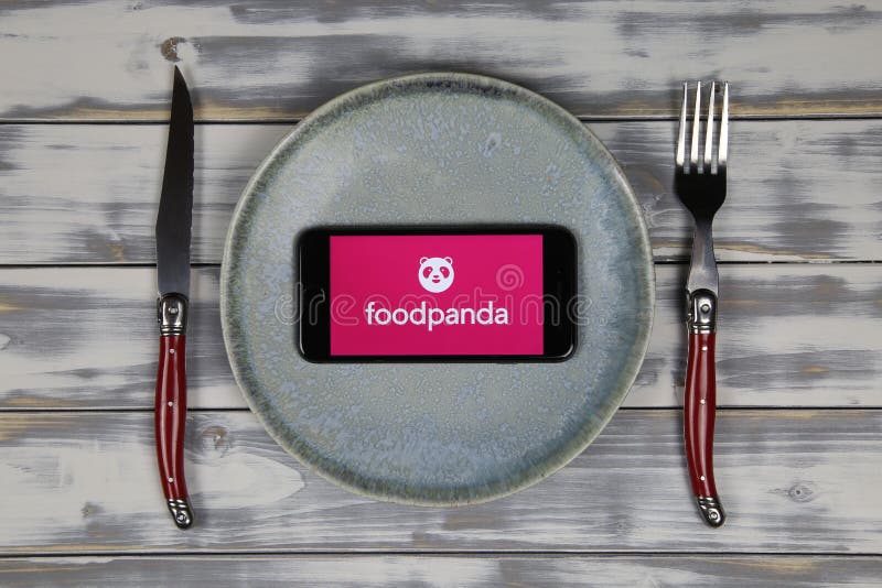 Viersen, Germany - May 9. 2020: Close up of mobile phone screen with logo lettering of food delivery service foodpanda on wood table with dish and cutlery focus on center